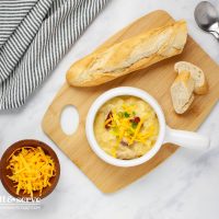 Crock of cheesy potato soup with kielbasa pieces garnished with shredded cheddar and sliced green onion on a cutting board with baguette slices beside bowls of sliced green onion and grated cheddar cheese.