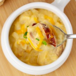 Closeup of a spoonful of potato soup with kielbasa garnished with shredded cheddar cheese and sliced green onion held above a crock of the soup.