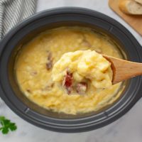 Close-up of a spoonful of cheesy potato soup with kielbasa pieces on a wooden spoon held over a crockpot of the soup.