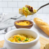 Crock of cheesy potato soup garnished with shredded cheddar cheese and sliced green onion with a spoonful held above the crock with a baguette in the background.