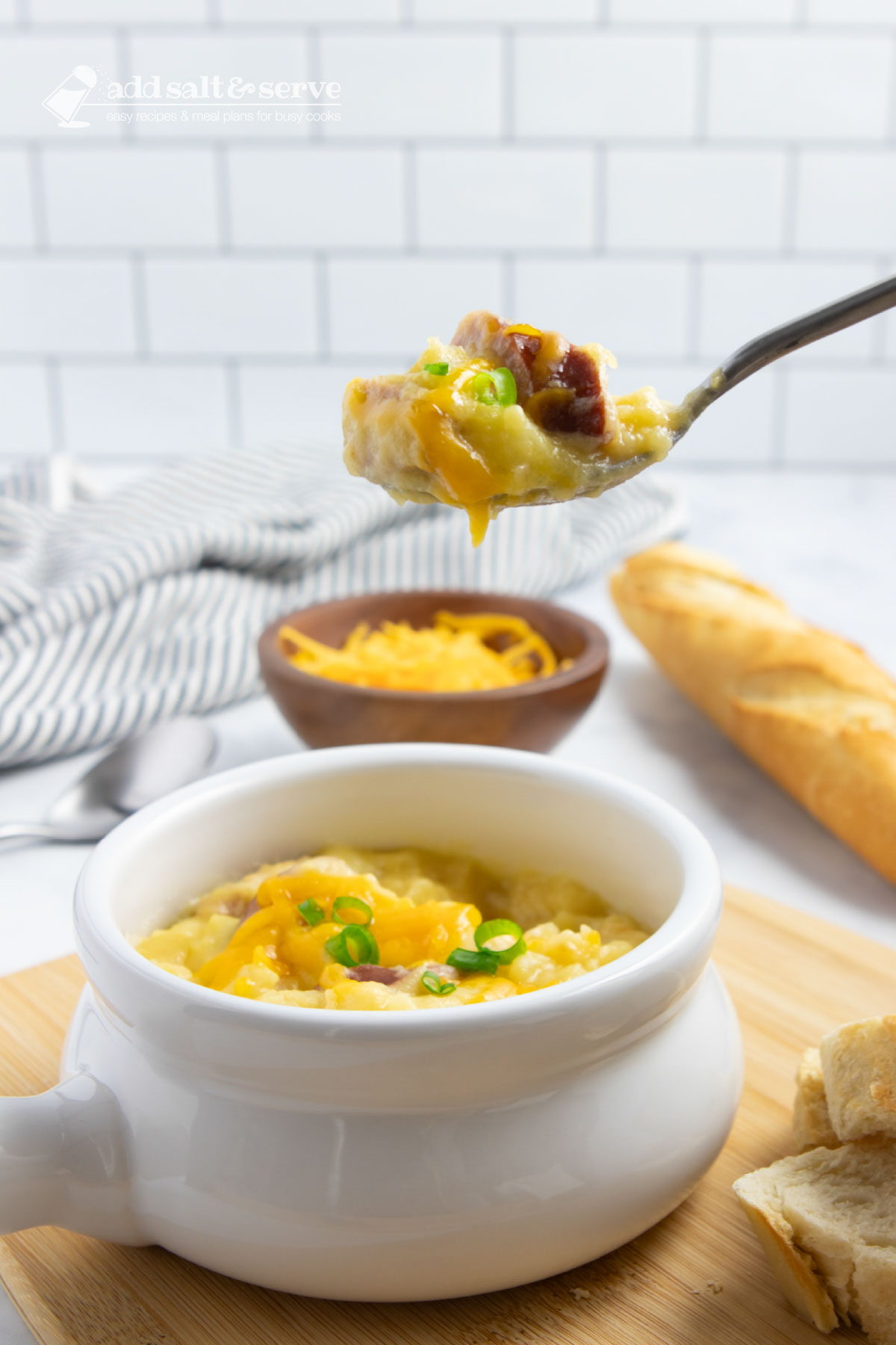 Crock of cheesy potato soup garnished with shredded cheddar cheese and sliced green onion with a spoonful held above the crock with a baguette in the background.