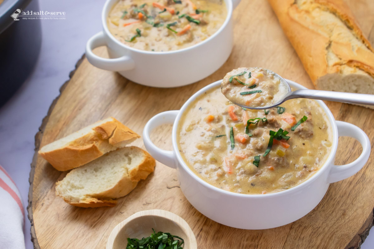 Two bowls of hash brown soup in white crocks garnished with fresh basil strips beside basil strips in a small bowl and slices of fresh baguette.