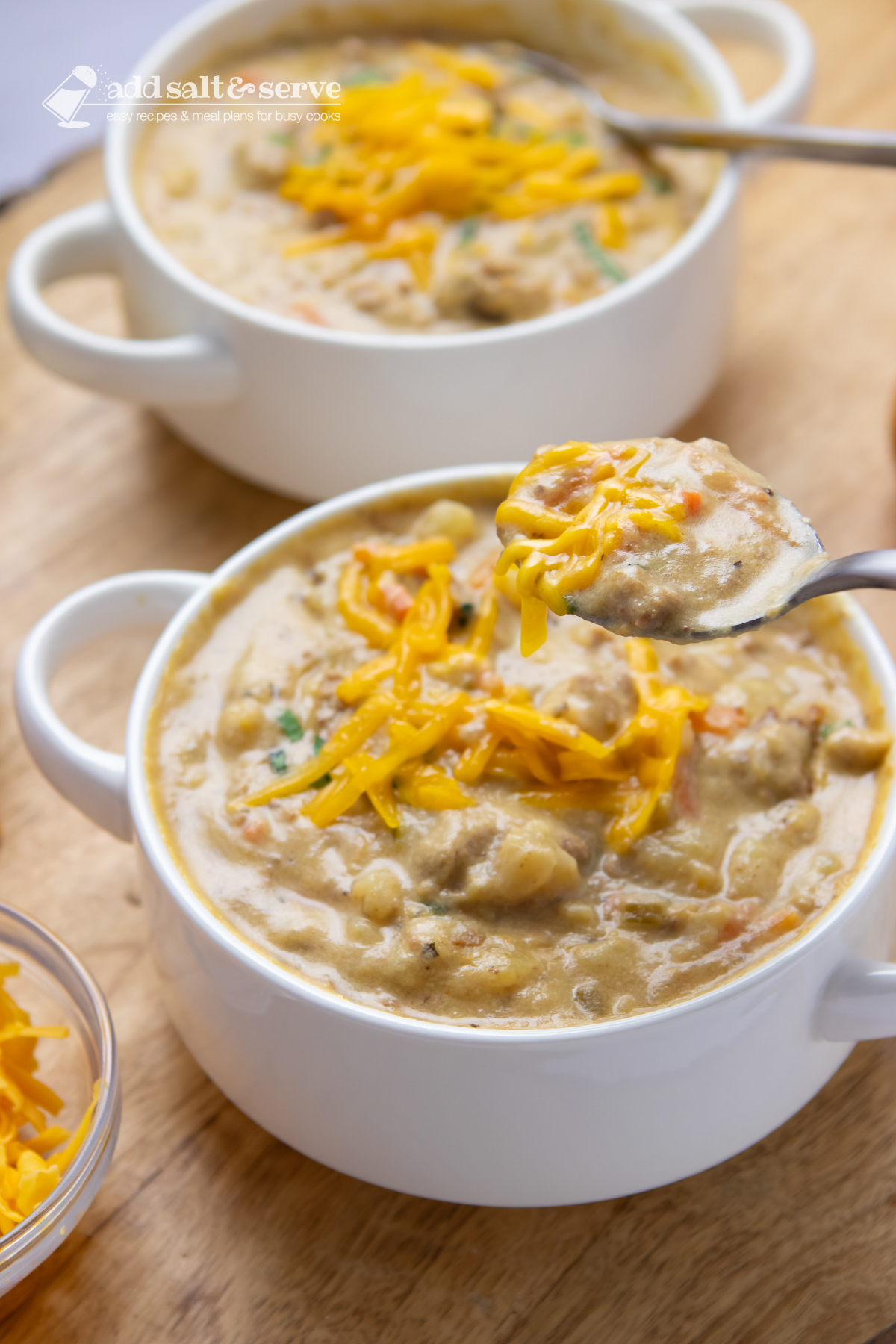 Two bowls of hash brown soup in white crocks garnished with shredded cheddar cheese and fresh basil strips with a closeup of a spoonful of the soup lifted above the bowl.