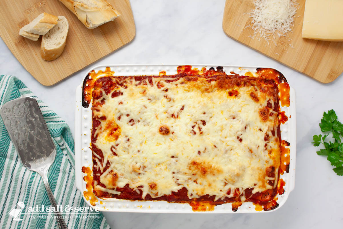 Pan of cheesy baked ziti on a counter surrounded by sprigs of fresh parsley, sliced baguette, and Parmesan cheese.