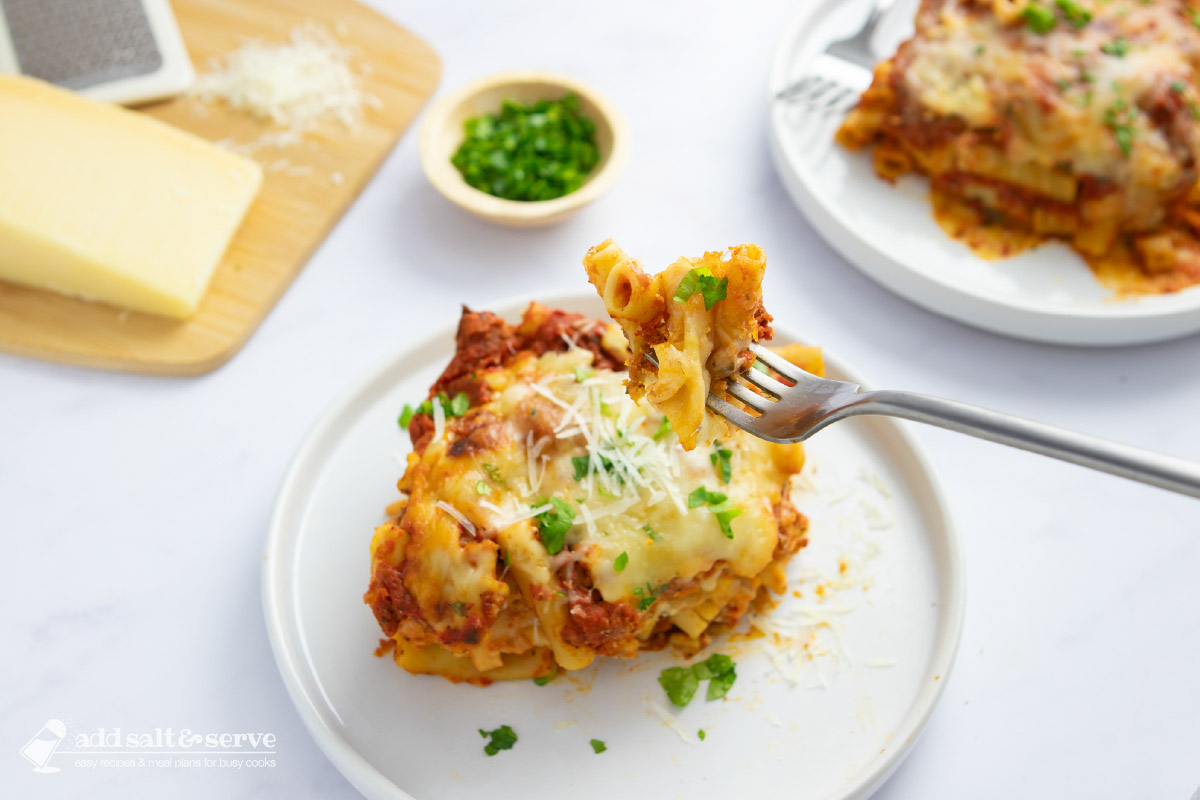 Two servings of cheesy meatless baked ziti in bowls beside Parmesan cheese on a cutting board with the baked ziti pan in the background.