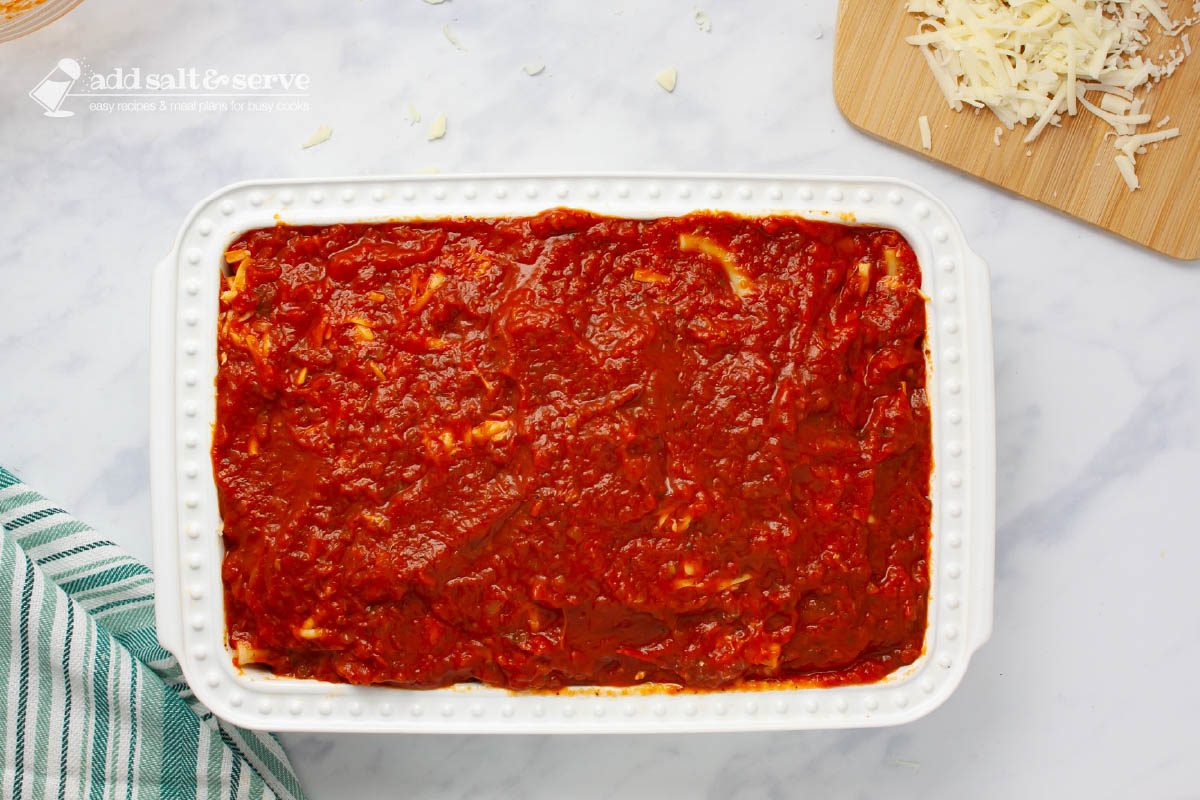 Pan showing a layer of marinara sauce spread over other layers of baked ziti.