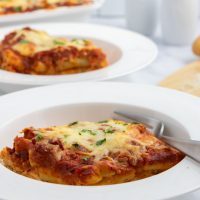 Two servings of cheesy meatless baked ziti in bowls beside Parmesan cheese on a cutting board with the baked ziti pan in the background.