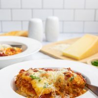 A serving of cheesy meatless baked ziti in a bowl with a fork with a cutting board with parmesan cheese in the background.