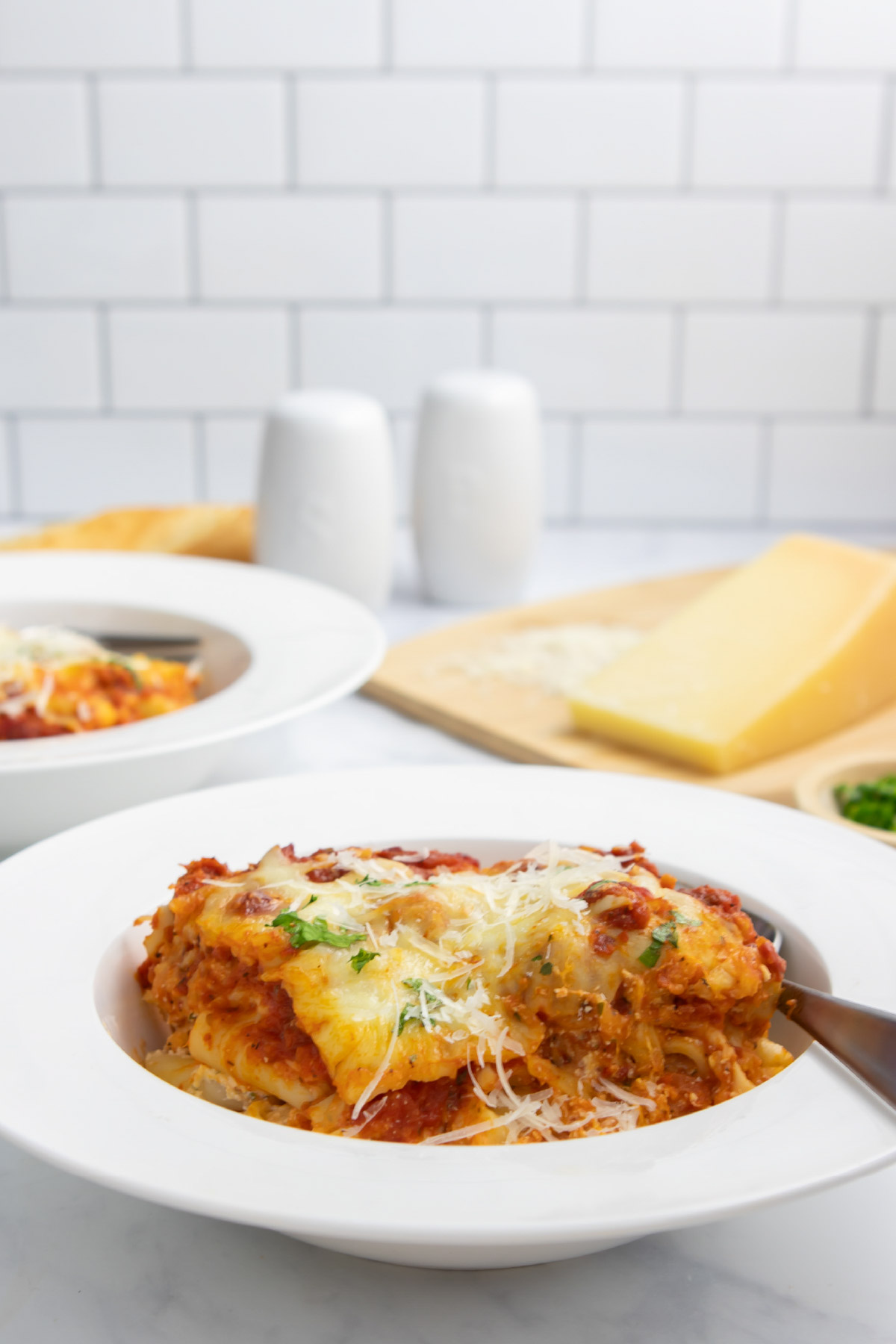 A serving of cheesy meatless baked ziti in a bowl with a fork with a cutting board with parmesan cheese in the background.