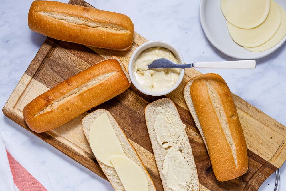 Hoagie rolls sliced lengthwise on a cutting board with provolone cheese on one side and horseradish sauce on the other side.